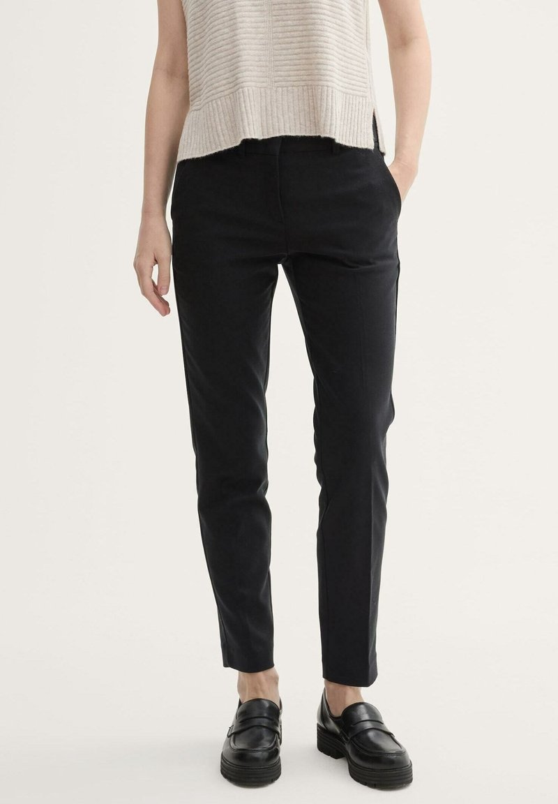 Personne portant un pantalon noir slim avec poches latérales, un haut beige clair sans manches et des chaussures compensées noires à enfiler.