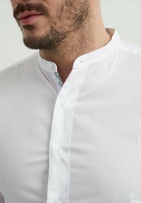 Chemise blanche à col classique avec un devant à boutons, présentant un motif floral à l'intérieur du col. Texture lisse et coupe ajustée.