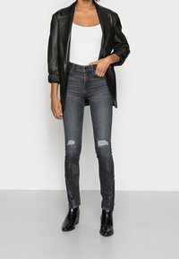 Blazer de cuero negro sobre una camiseta blanca, combinado con jeans ajustados gris oscuro con detalles rasgados y botas negras con acabado texturizado.