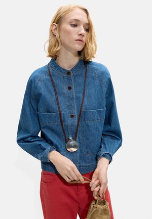 Blondynka w niebieskiej jeansowej kurtce z brązowymi guzikami, długim naszyjnikiem z srebrnym wisiorkiem i czerwonymi spodniami, trzymająca beżową zamszową torbę.