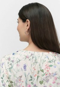 Blomstret bluse med en lys cremefarvet baggrund, der har flerfarvede blomstermønstre. Bagsidedesignet inkluderer en lille cirkulær knapdetalje.