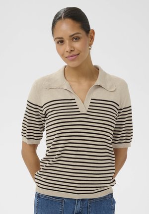 Nő, aki bézs színű, fekete vízszintes csíkos, rövid ujjú pulóvert visel nyitott gallérral, kezeit a hát mögött tartja, enyhén mosolyog.