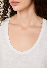 Balta, lengva marškinėliai su plačiu apvaliu iškirpimu, pagaminti iš minkštos, tekstūruotos medžiagos ir turintys subtilų melanzavimo raštą krūtinės srityje.