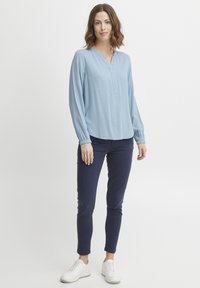 Világoskék, hosszú ujjú blúz V-nyakkal és gumírozott mandzsettákkal, sötétkék, slim-fit nadrággal és fehér sportcipővel párosítva.