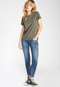 T-shirt en coton vert olive à manches courtes avec un imprimé graphique. Associée à un jean slim bleu et des baskets montantes noires.