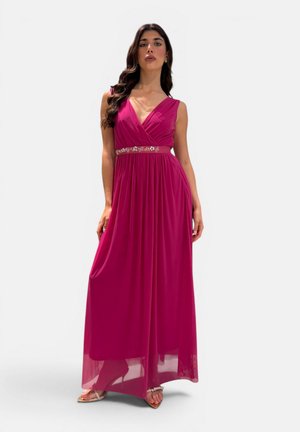 Donna che indossa un abito lungo senza maniche di colore rosa intenso con scollo a V e cintura floreale, in piedi con una mano che tiene l'orlo dell'abito.