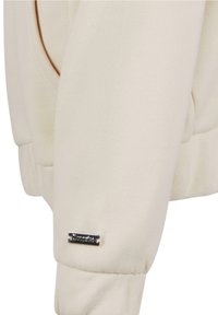 Gros plan sur la manche d'un sweat-shirt beige avec une petite étiquette métallique rectangulaire de marque près du poignet.