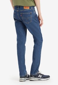 Blå denim bukser med rett passform, med baklommer, et skinnmerke og teksturert stoff. Kombinert med mørke sportsko.