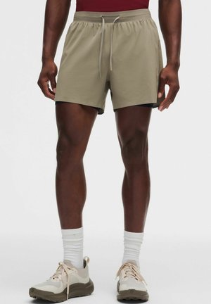 Hombre con pantalones cortos deportivos beige con cordón, calcetines blancos y zapatillas de correr beige, de pie frente a un fondo liso.