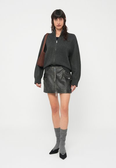 Casaco de malha cinzento escuro com fecho éclair, minissaia de couro preta com fecho frontal, meias cinzentas caneladas e saltos agulha pretos, tudo sobre um fundo neutro.