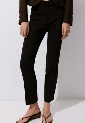 Personne portant un pantalon noir ajusté longueur cheville, une veste courte marron foncé, et des sandales compensées marron avec des bagues aux deux mains.