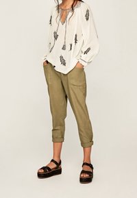 Weiße Bluse mit schwarzen Blattmustern und Quastendetails, kombiniert mit olivgrünen hochgekrempelten Hosen und schwarzen Plateausandalen.