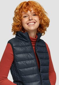 Granatowa pikowana kamizelka puchowa z wysokim kołnierzem, zamkiem błyskawicznym i gładką teksturą, noszona na pomarańczowej koszuli z długim rękawem.