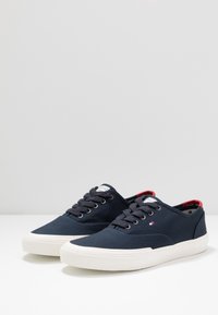 Marineblaue Canvas-Sneaker mit weißen Gummisohlen, runder Zehenpartie, strukturiertem Obermaterial, Schnürdesign und roten Akzenten an der Ferse und am inneren Kragen.