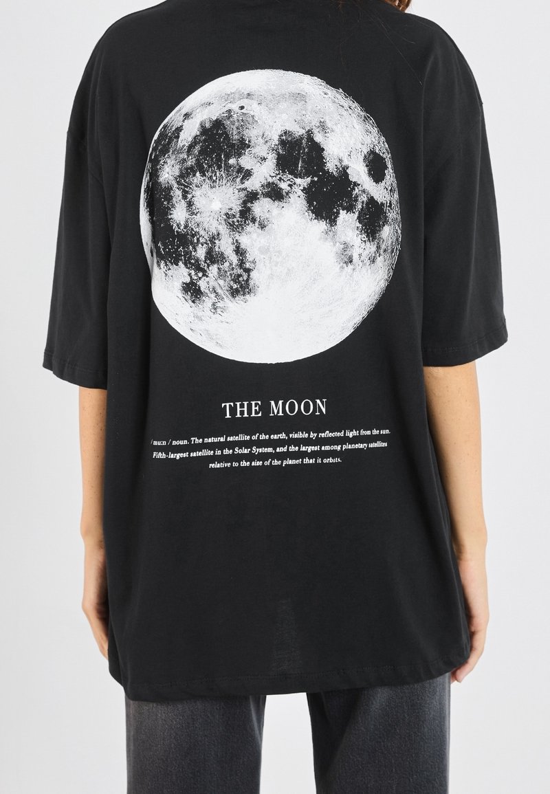 Sort oversized t-shirt med en stor hvid afbildning af månen på ryggen, med tekst under grafikken. Enkel, blød bomuldsstof.