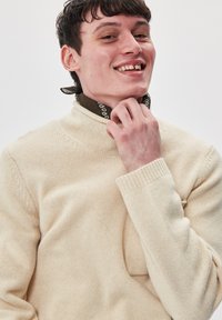 Cremfarbener gestrickter Pullover mit hohem Kragen, gerippten Bündchen und sichtbarer Textur. Ein gemusterter brauner Schal liegt um den Hals.