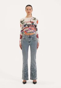 Langsleeve, doorschijnende crop top met kleurrijke bloemenprints en de tekst "LOVE HARD." Gecombineerd met lichtgekleurde jeans met een textuurpatroon.