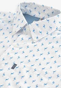 Camisa blanca con un patrón repetido de flamencos azules, cuello abotonado y un bolsillo con una pequeña etiqueta de logo.