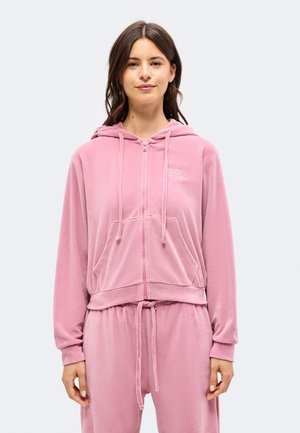 Femme portant un sweat à capuche rose zippé avec poches avant et pantalon rose à cordon assorti, debout devant un fond clair uni.