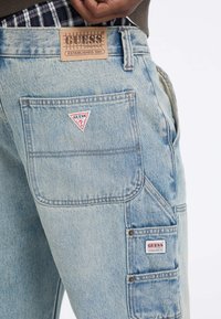 Shorts cargo in denim azzurro chiaro con multiple tasche, caratterizzati da patch con il logo Guess sulle tasche posteriori e laterali. Design strutturato e cuciture definite.