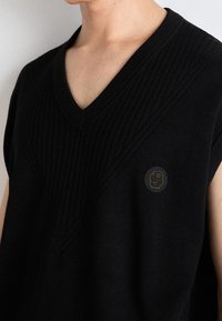 Czarny, dziergany bezrękawnik z dekoltem w serek, z teksturą prążkowaną oraz okrągłym logo z literą "B" w złotym kolorze po lewej stronie.