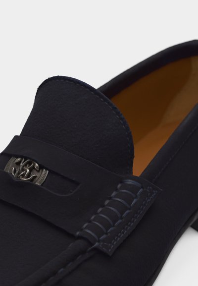 Κοντινό πλάνο ενός μαύρου δερμάτινου μοκασίνι με λεπτομερείς ραφές και ασημένιο διακοσμητικό μεταλλικό έμβλημα στο λουράκι.