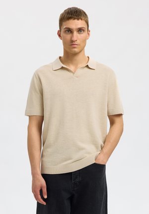 Polo de punto color beige claro con una textura suave, mangas cortas y cuello con muesca; combinado con pantalones negros.
