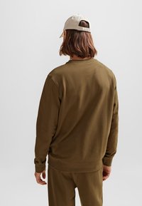 Felpa verde oliva con scollatura a girocollo, maniche lunghe e polsini a costine, abbinata a un cappellino beige. Il tessuto sembra morbido e strutturato.