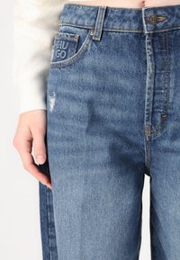 Zils denims ar brīvu piegriezumu, redzamām šuvēm, nelielām nolietojuma pazīmēm un birku ar uzrakstu "HUGO". Pogas aizdare jostas vietā.