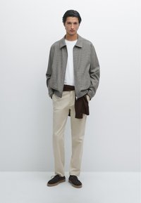 Giacca bomber grigia con texture sopra una maglietta bianca, abbinata a pantaloni beige e scarpe marroni, con un maglione marrone annodato in vita.