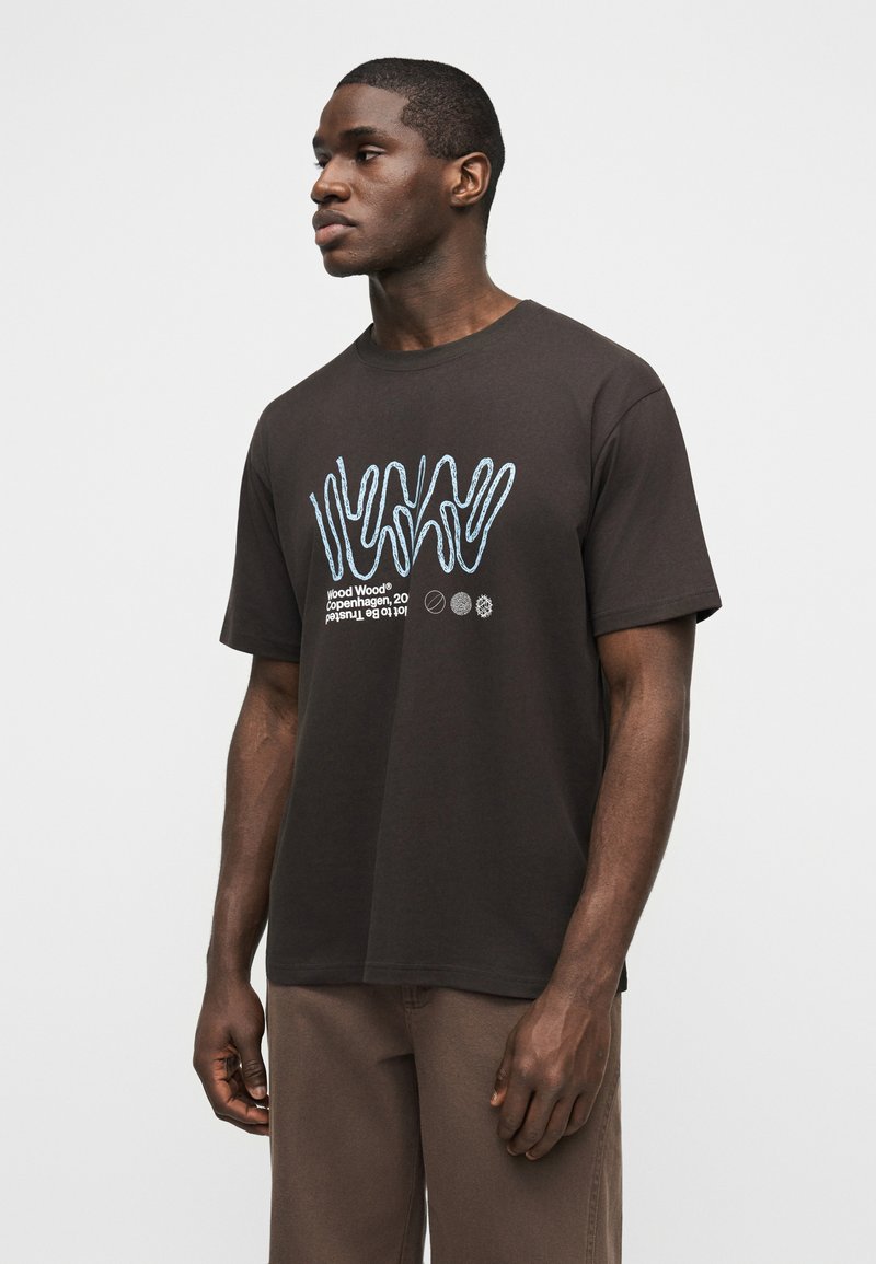 Young Black man wearing a black T-shirt with blue abstract lines and white text, standing against a plain light background.