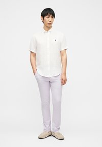 Άνδρας που στέκεται με τα χέρια στις τσέπες, φορώντας λευκό πουκάμισο με κοντά μανίκια και κουμπιά, ανοιχτόχρωμο λιλά παντελόνι και μπεζ μοκασίνια.