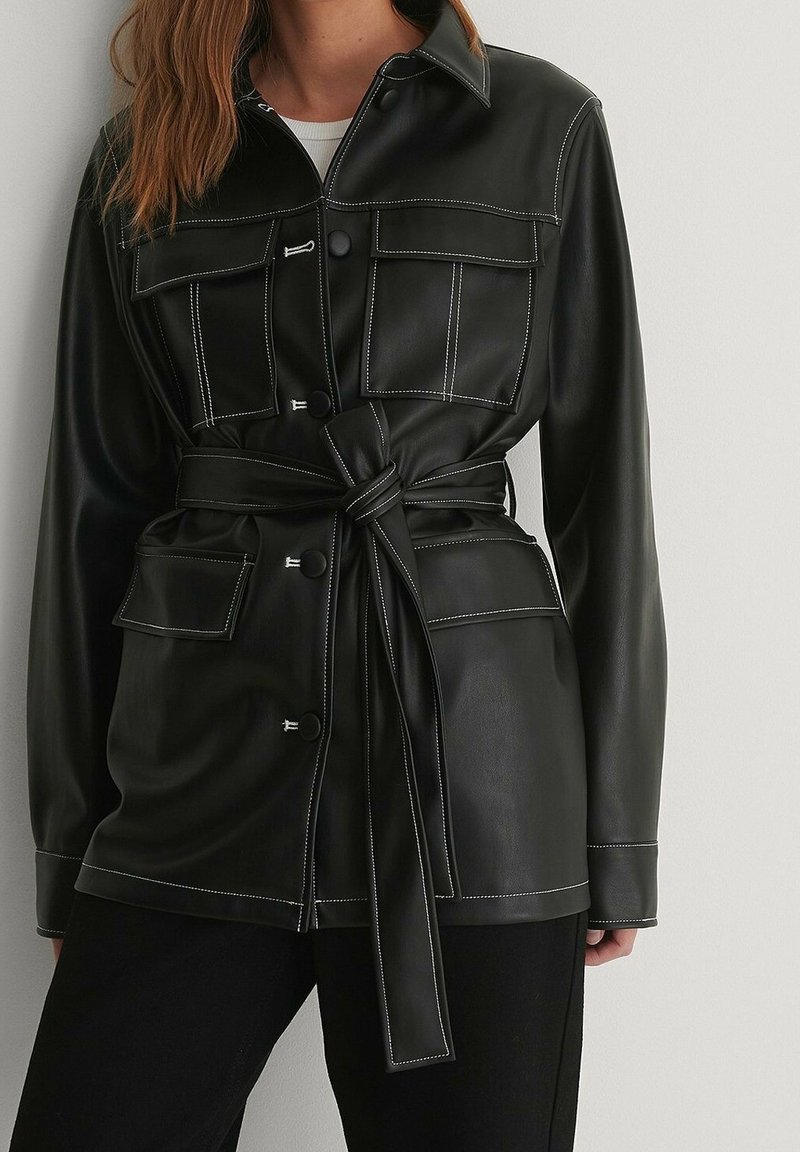 Chaqueta de cuero sintético negra con dos bolsillos en el pecho, cintura con cinturón, costuras blancas en contraste y un diseño frontal con botones.