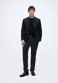 Uomo in piedi su sfondo bianco che indossa un abito blu scuro, camicia nera, cintura nera e scarpe nere, con una mano in tasca.
