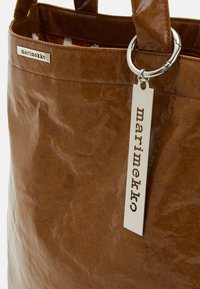 Sac en papier brun avec une texture brillante, présentant un anneau en argent et une étiquette avec le texte "marimekko". Les poignées sont robustes et fixées solidement.