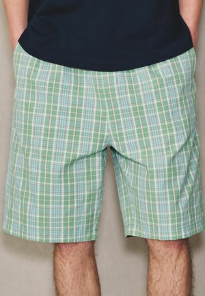 Mann trägt grüne und weiße karierte Shorts und ein schwarzes Hemd, die Hände in den Taschen, vor neutralem Hintergrund.