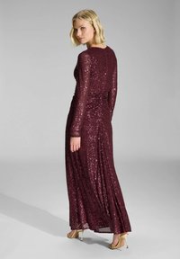 Robe longue en sequins bordeaux avec manches longues et une silhouette ajustée. Présente un ruching à la taille, associée à des chaussures à talons hauts dorées.