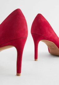 Czerwone zamszowe szpilki o spiczastym nosku i smukłym obcasie typu stiletto. Tekstura jest miękka, podkreślająca gładkie wykończenie.