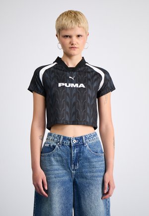 Mlad oseba s kratkimi blond lasmi, ki nosi obročne uhane, črno krojeno športno majico Puma in široke izbledele modre kavbojke, stoji ob belem ozadju.