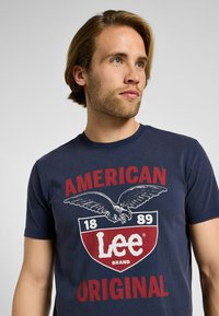 Camiseta de color azul marino hecha de algodón con cuello redondo. Presenta un diseño gráfico audaz con un águila, la palabra "Lee" y elementos de texto vintage.