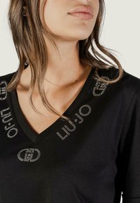 Czarna koszulka V-neck z miękkiego materiału, ozdobiona kryształowymi akcentami oraz logo marki wzdłuż dekoltu. Zbliżenie od ramion w górę.