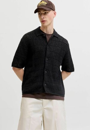 Mladý muž s hnedou čiapkou, čiernou krátkym rukávom háčkovanou košeľou na gombíky cez hnedé tričko a svetlými nohavicami, stojaci s rukami za chrbtom.