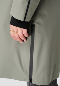 Main avec des bagues en or près d'un zip noir sur la manche d'un manteau gris clair superposé sur un poignet noir contre un fond blanc.