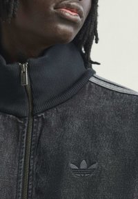 Črn jeans jakna z rebrastim črnim ovratnikom, srebrnim zadrgo in diskretnim črnim logotipom na prsih. Na ramah so tri sive črte.