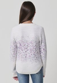 Frau mit langen braunen Haaren, die einen weißen Pullover mit lila gepunktetem Muster und blaue Jeans trägt, von hinten vor einem schlichten Hintergrund gezeigt.