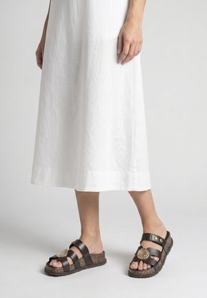 Piernas bajas con sandalias marrones tipo slip-on con medallones decorativos, combinadas con una falda blanca hasta el tobillo.