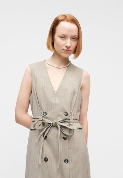 Νέα γυναίκα με κοντά κόκκινα μαλλιά, φορώντας μπεζ αμάνικο φόρεμα με κουμπιά και ζώνη στη μέση, καθώς και ένα κολιέ με πέρλες, στέκεται μπροστά σε λευκό φόντο.