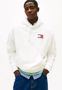 Sudadera blanca de algodón con cordón, con un logotipo gráfico en púrpura y rojo. Combinada con una camiseta de rayas verdes y azules debajo.