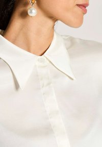 Witte blouse met een klassieke kraag, met een knoopsluiting bij de hals. De stof is glad en lichtgewicht. Pareloorbel accent.