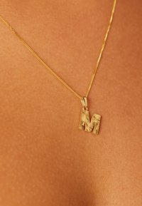 Gouden ketting met een gestructureerde hanger in de vorm van de letter "M." De ketting is fijn en delicaat en rust tegen een huidkleurige achtergrond.